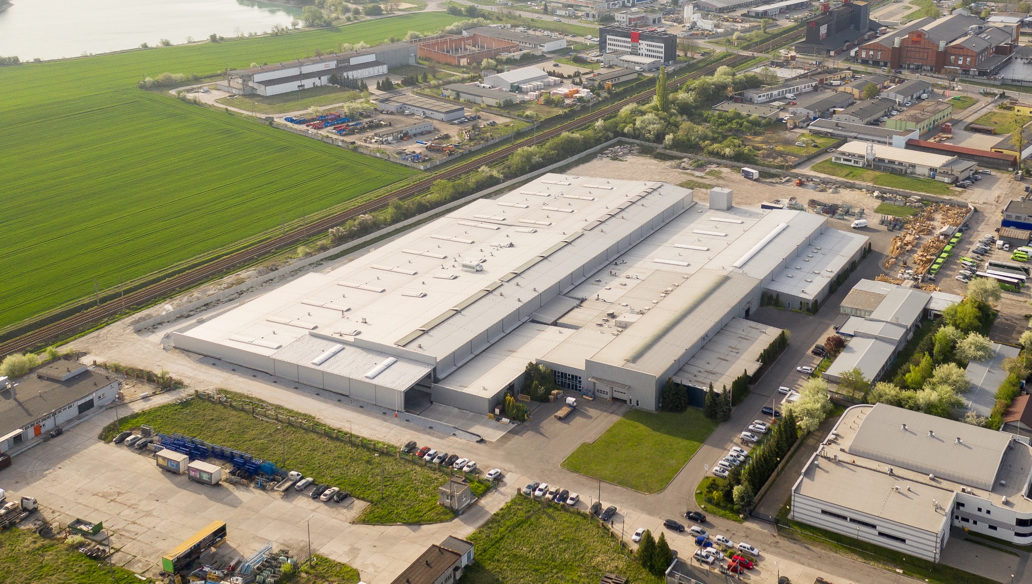 Szeroki kadr z lotu ptaka przedstawiający kompleks hal przemysłowych z blachy trapezowej, otoczonych zielenią pól i zabudową miejską.