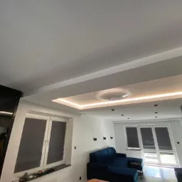 Nowoczesne wnętrze salonu z podwieszanym sufitem z oświetleniem LED, widoczna część niebieskiej sofy oraz okna z roletami.