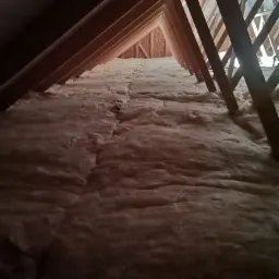 Izolacja z wełny mineralnej między krokwiami poddasza, widok z dołu, drewniana konstrukcja dachu z widocznym niebieskim niebem przez szczeliny.