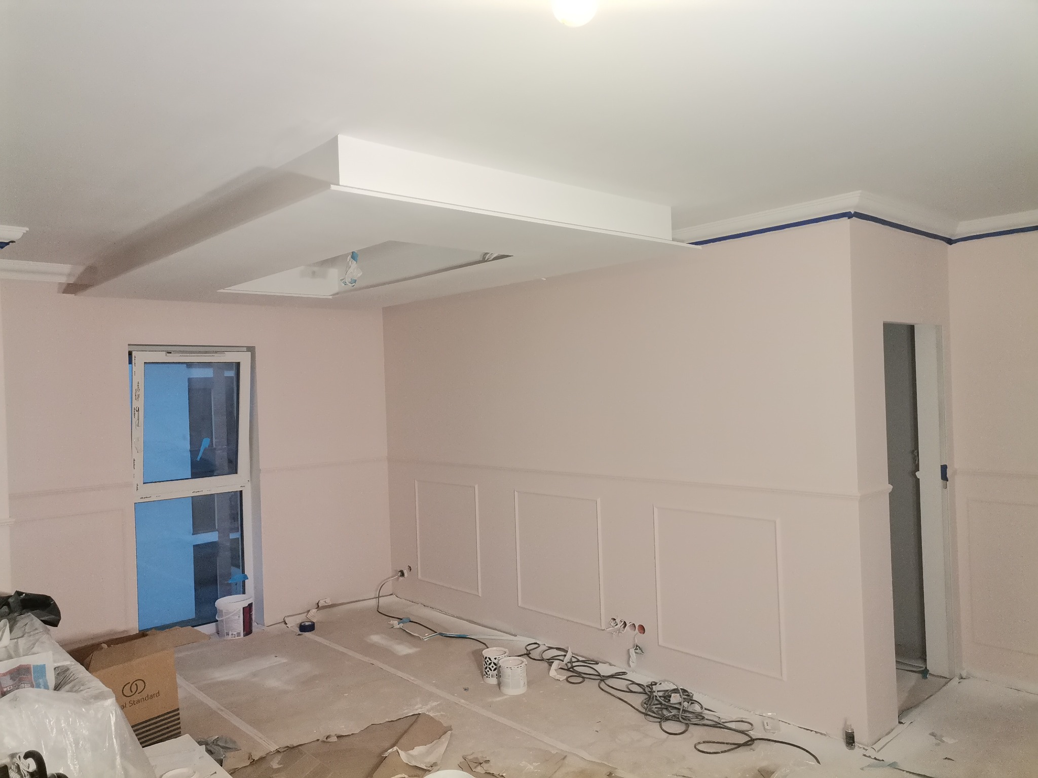 Wnętrze w trakcie prac wykończeniowych: różowe ściany z dekoracyjnymi panelami, zabezpieczone taśmą malarską listwy przysufitowe, podłoga pokryta warstwą pyłu, widoczne puszki z farbą i narzędzia...