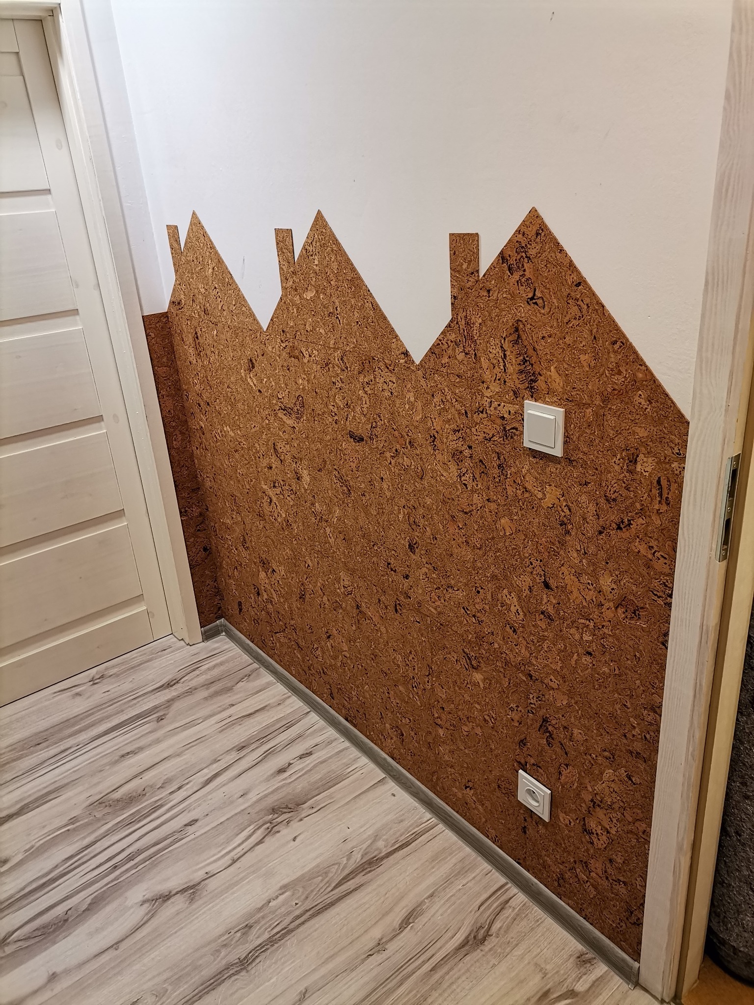 Kreatywna aranżacja ściany z korka w kształcie domków, z widocznym włącznikiem światła i gniazdkiem elektrycznym, na tle jasnej podłogi imitującej drewno.