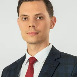 Portret młodego mężczyzny w ciemnym garniturze w kratę i czerwonym krawacie, na białym tle.