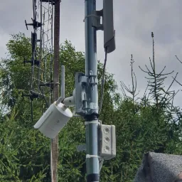 Zewnętrzna kamera monitoringu Dahua zamontowana na metalowym słupku obok anteny telewizyjnej, na tle zielonych drzew i pochmurnego nieba.