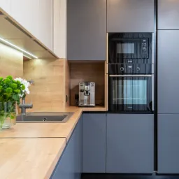 Nowoczesna kuchnia z szarymi szafkami i drewnianym blatem, z wbudowanym piekarnikiem i mikrofalówką marki Gorenje, ekspres do kawy w niszy, zielone kwiaty w wazonie przy zlewie, oświetlenie LED...