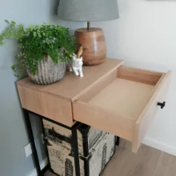 Drewniana szafka nocna z wysuwaną szufladą, stojąca na metalowym stelażu z motywem podróżniczym, na blacie doniczka z paprocią, lampka i figurka renifera.