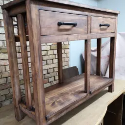 Drewniana konsola z dwiema szufladami i dolną półką, wykończona w ciepłym brązie, ustawiona na blacie roboczym z ceglanym murem w tle.