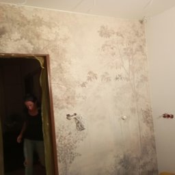 Szybki Remoncik - Ściana z tapetą w odcieniach szarości przedstawiającą motyw leśny, widoczne niedokończone gniazdka elektryczne i fragment osoby w drzwiach.