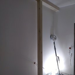 Szybki Remoncik - Szlifowanie ściany szlifierką z oświetleniem halogenowym w trakcie remontu, widoczne kable elektryczne i drewniana konstrukcja na ścianie.
