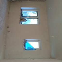 Szybki Remoncik - Wnętrze pokoju w trakcie remontu: ściany po nałożeniu gładzi, zabezpieczone okna folią, widoczny surowy sufit z cegły i wystające kable.