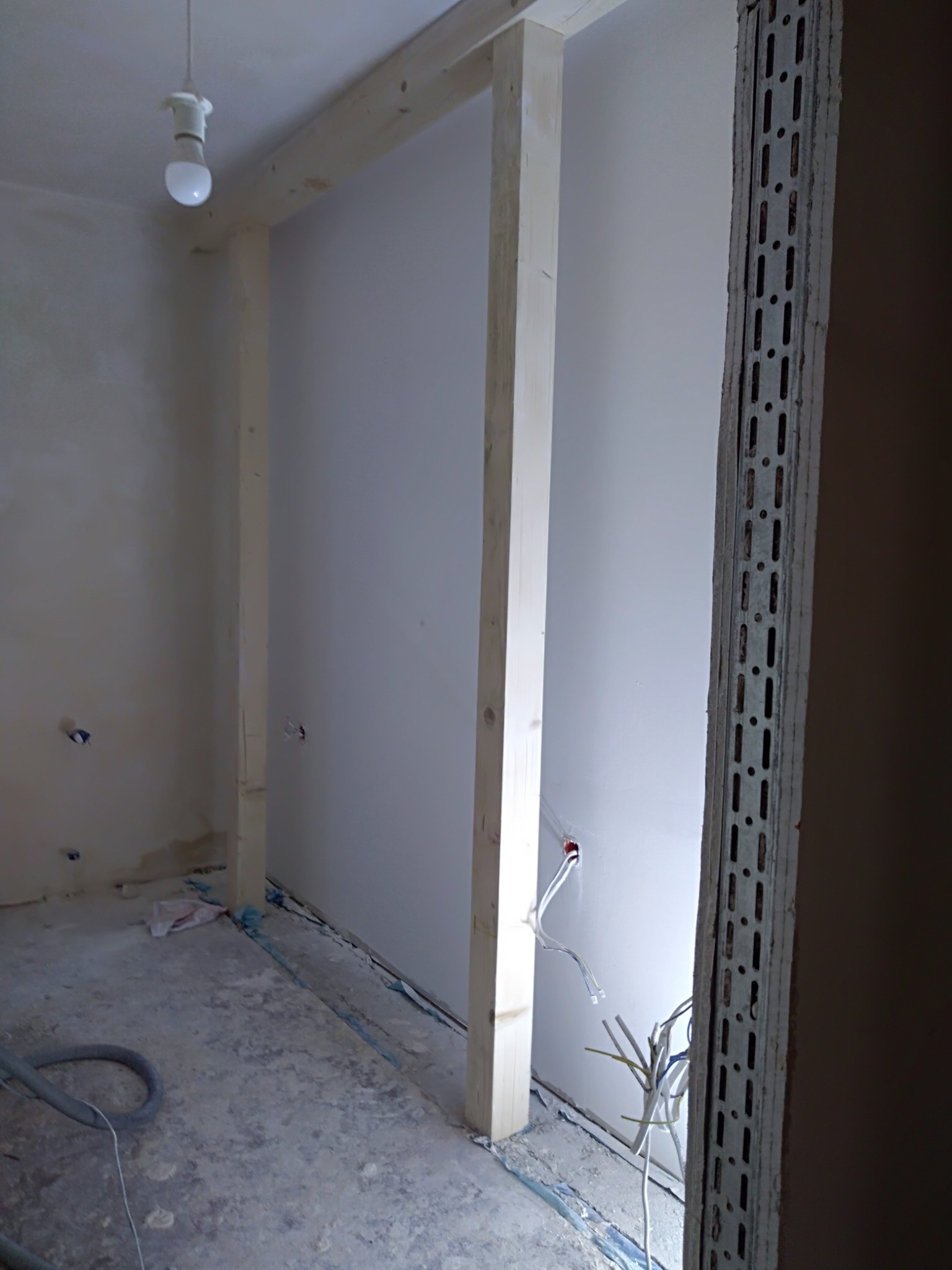 Wnętrze w trakcie remontu: drewniana konstrukcja ścianki działowej, metalowa listwa narożnikowa, wystające przewody elektryczne, surowy beton na podłodze, wisząca żarówka