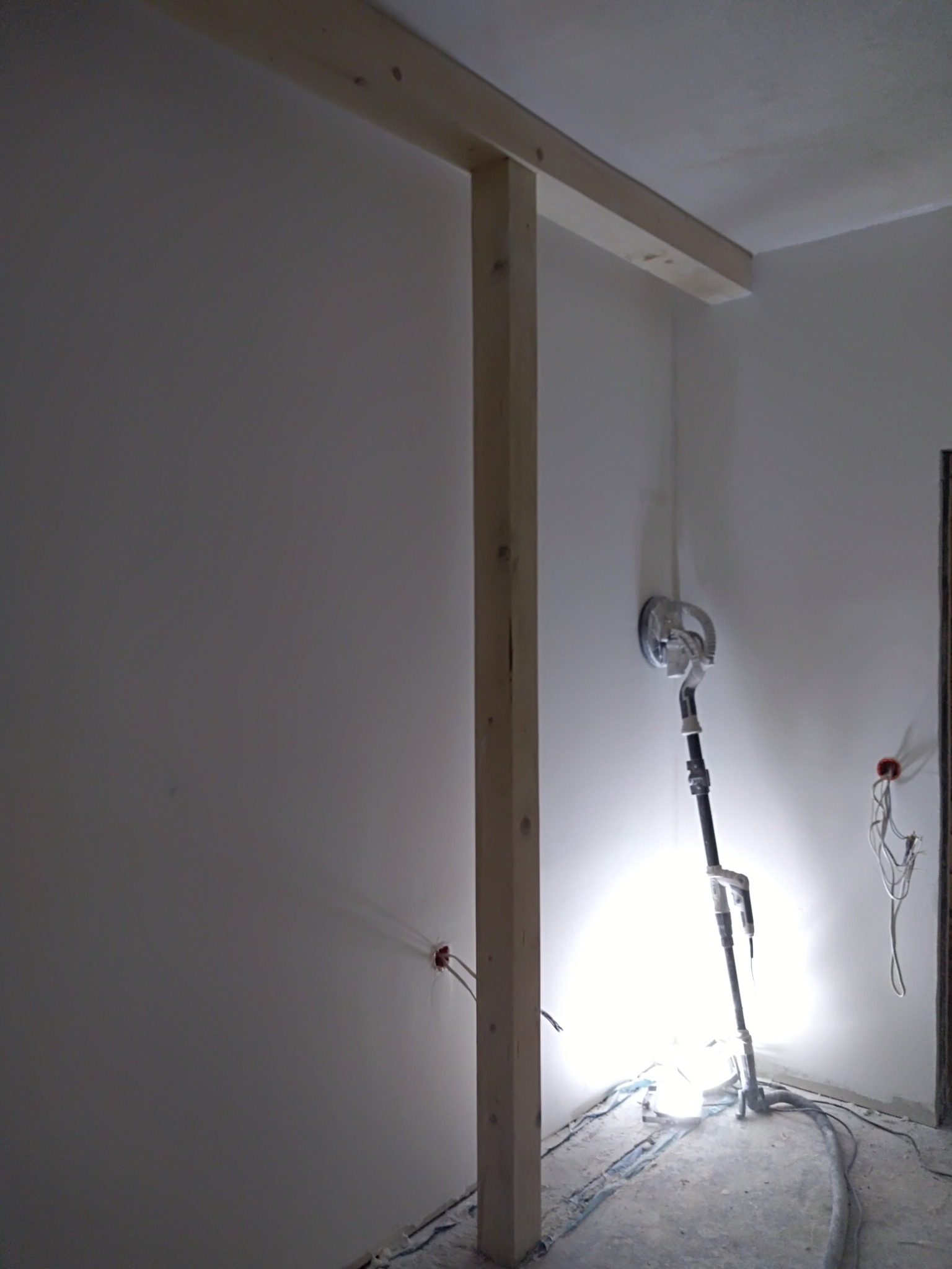 Szlifowanie ściany szlifierką z oświetleniem halogenowym w trakcie remontu, widoczne kable elektryczne i drewniana konstrukcja na ścianie.