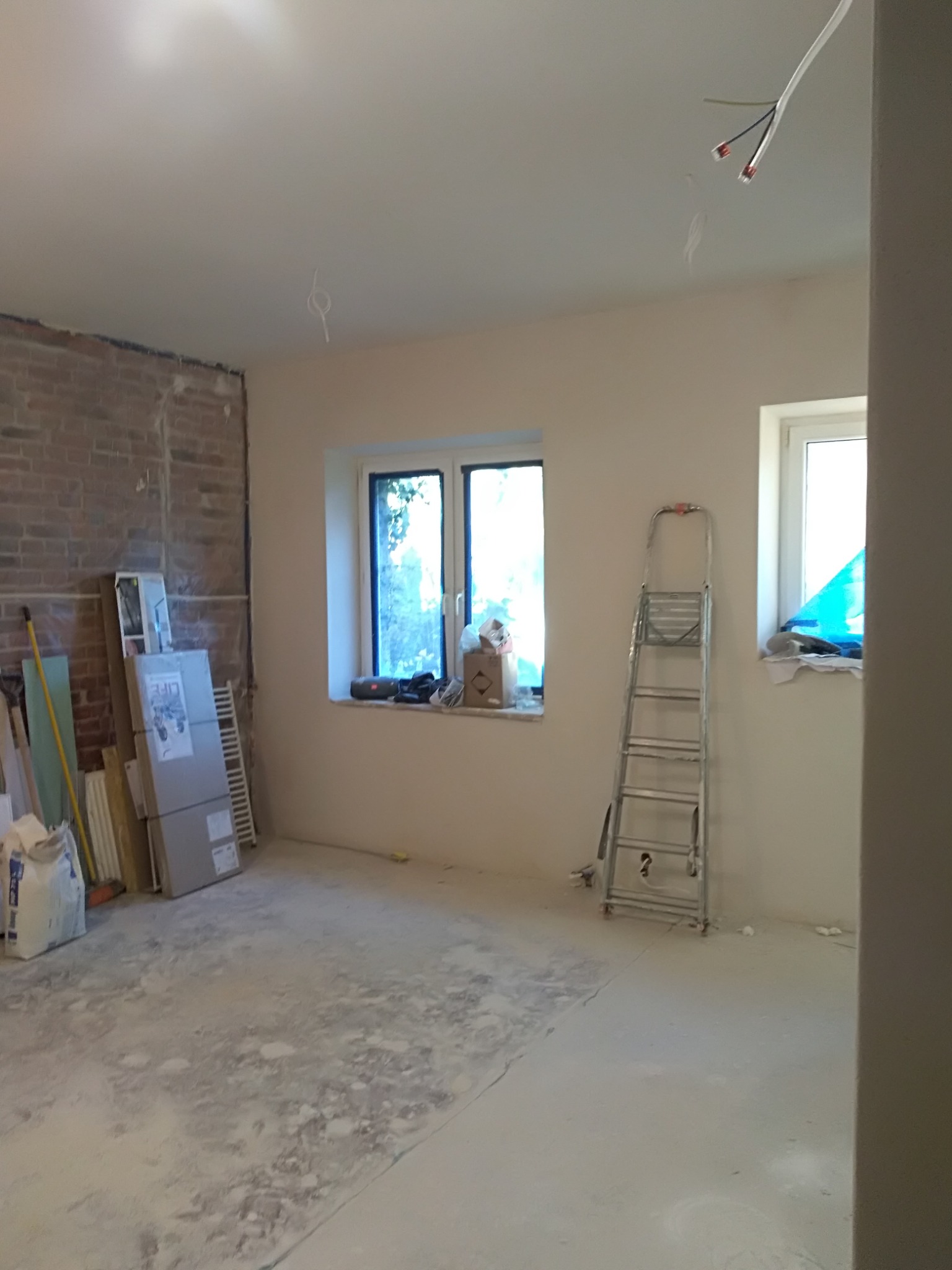 Pokój w trakcie remontu: odsłonięta cegła na jednej ścianie, przygotowane płyty gipsowe, drabina aluminiowa, surowe przewody elektryczne zwisające z sufitu.