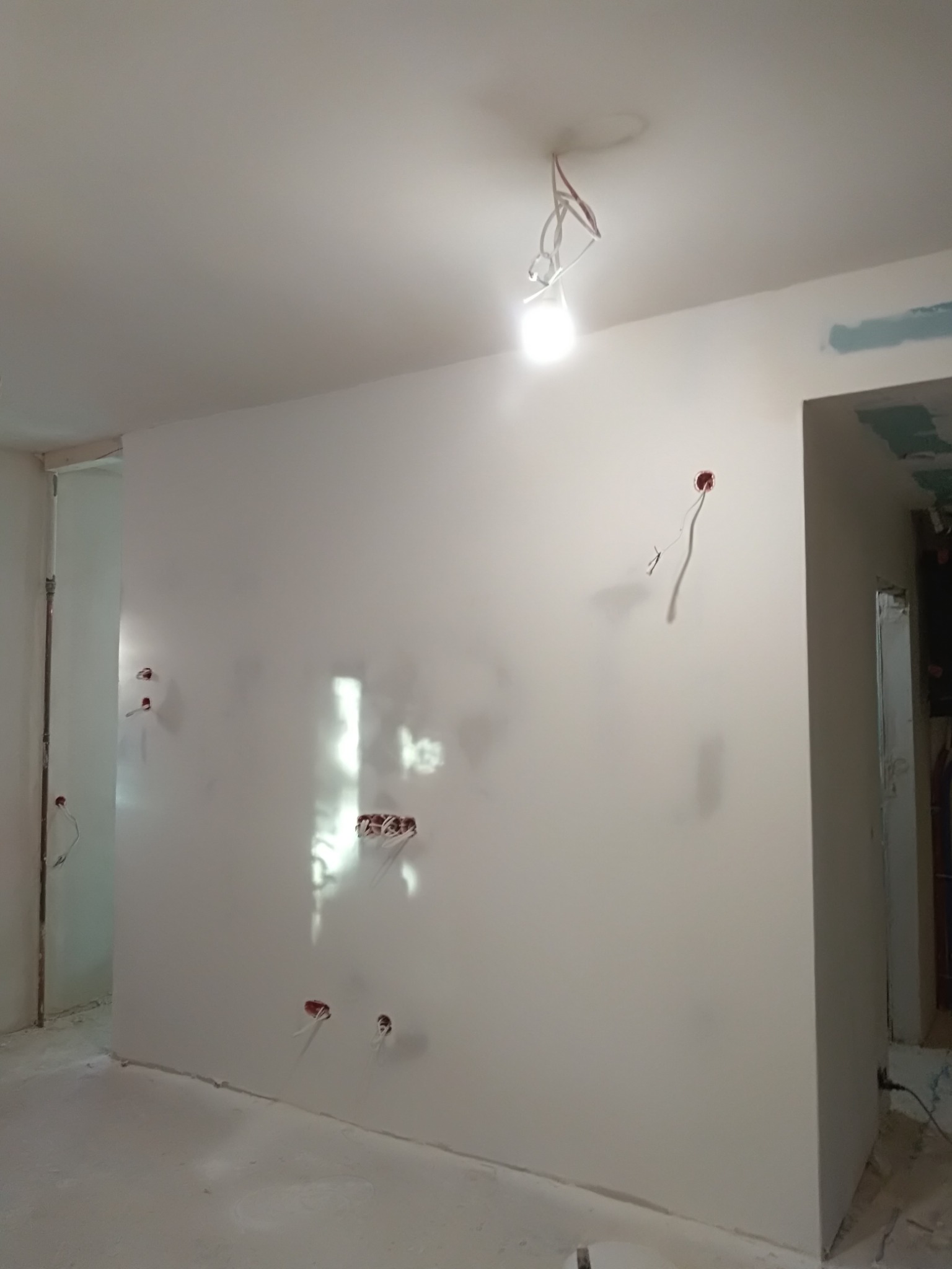 Wnętrze w trakcie remontu: świeżo wyszpachlowana ściana z widocznymi przewodami elektrycznymi, surowy sufit z prowizorycznym oświetleniem, przygotowane do dalszych prac wykończeniowych.