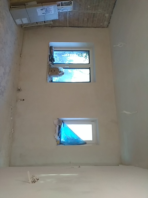 Wnętrze pokoju w trakcie remontu: ściany po nałożeniu gładzi, zabezpieczone okna folią, widoczny surowy sufit z cegły i wystające kable.