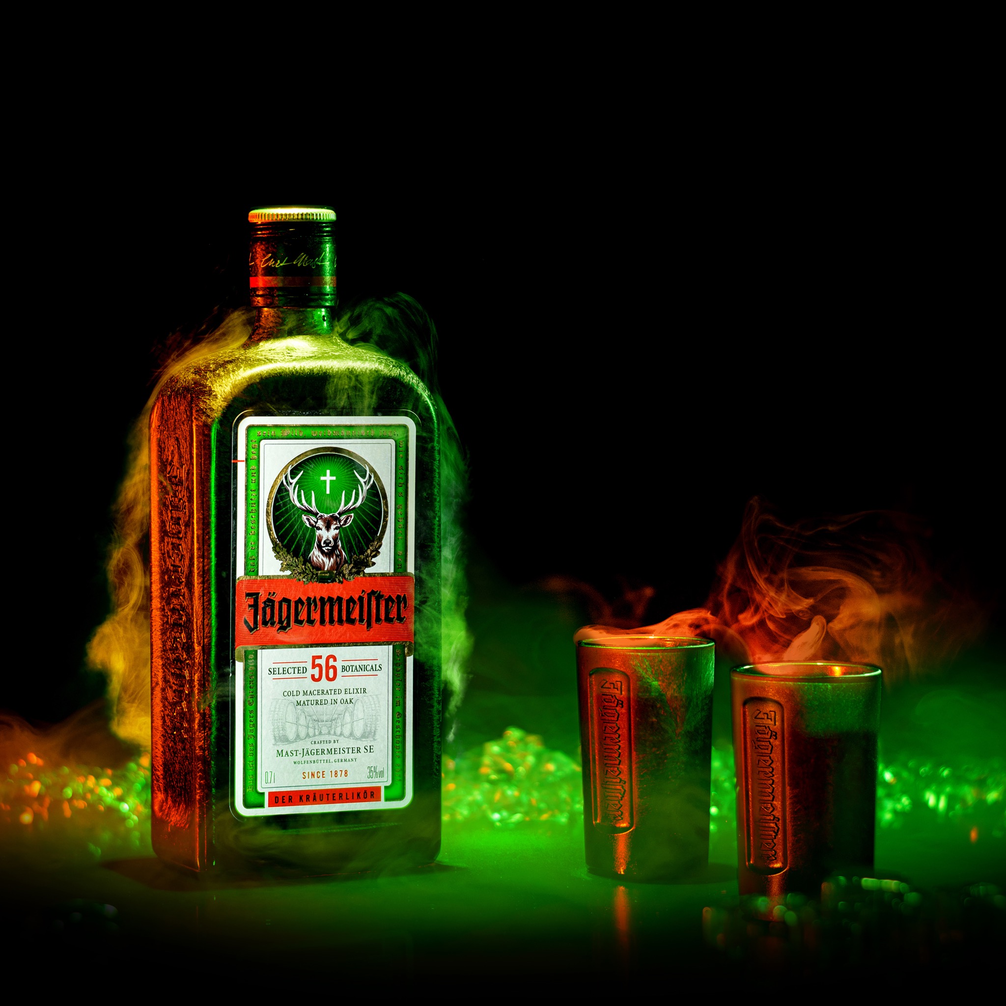 Butelka Jägermeistera i dwa metalowe kieliszki w oparach dymu, podświetlone zielonym i pomarańczowym światłem na czarnym tle.