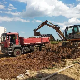 Czerwona wywrotka MAN i koparka CASE podczas prac ziemnych na budowie w słoneczny dzień, w tle dom jednorodzinny.