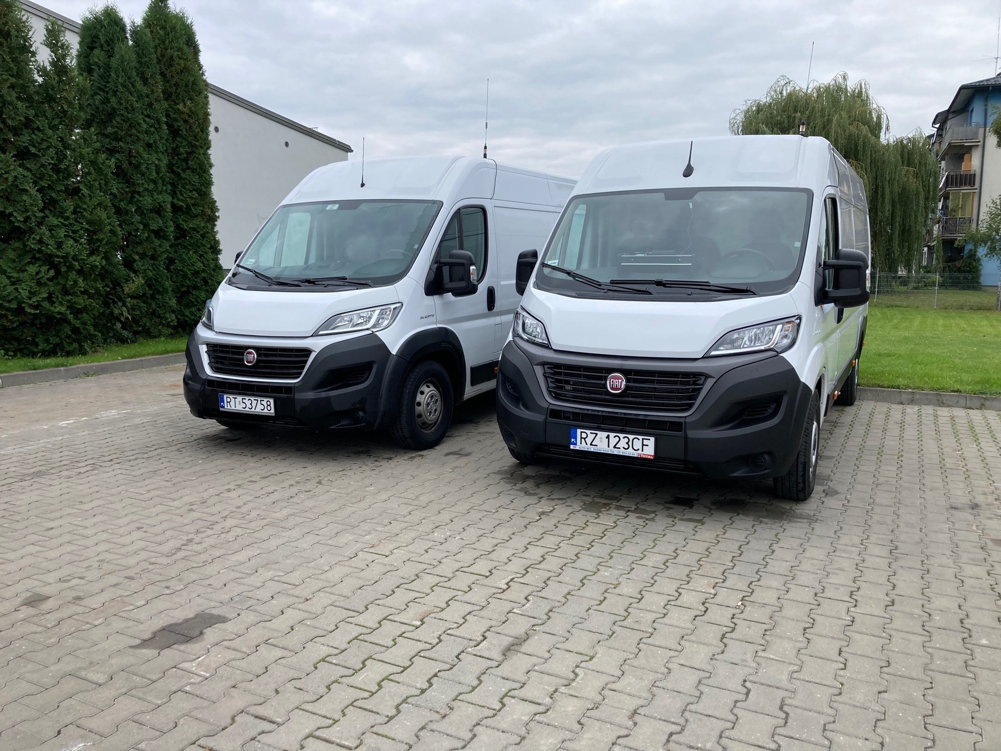 Dwa białe busy Fiat Ducato zaparkowane na brukowanym placu, z antenami na dachu, widoczne tablice rejestracyjne.
