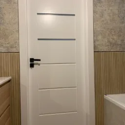 Nowoczesne, białe drzwi wewnętrzne z czarną klamką w łazience wykończonej płytkami w dwóch odcieniach beżu.