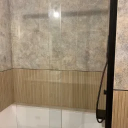 Kabina prysznicowa z przesuwnymi szklanymi drzwiami w łazience z płytkami w dwóch odcieniach beżu, widoczna bateria prysznicowa z deszczownicą.