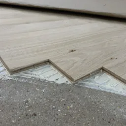 Fragment podłogi z jasnych, drewnianych paneli układanych na klej, widoczny surowy betonowy podkład.