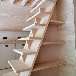 Surowe drewniane schody ażurowe z trójkątnymi wspornikami, umieszczone w pomieszczeniu wykończonym boazerią, z widocznymi elementami instalacji elektrycznej i zabudową meblową w tle.