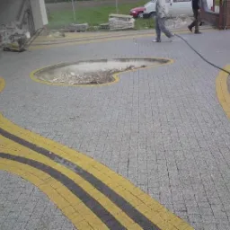 Kładzenie szarej kostki brukowej z żółtymi i czarnymi akcentami na podjeździe w Sadykierzu, widoczny fragment przygotowanego miejsca na rabatę.