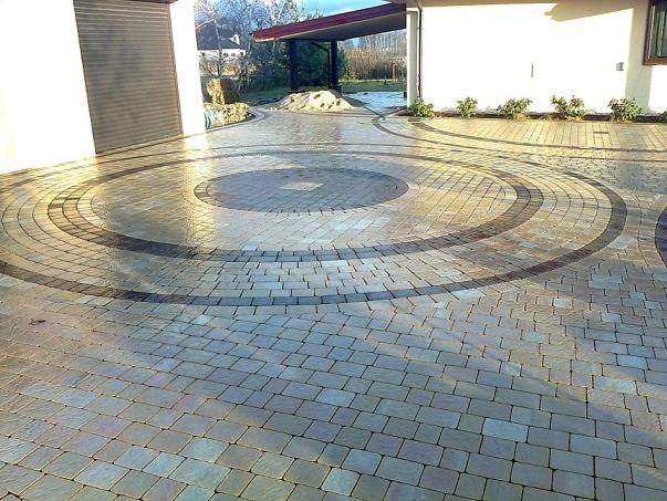 Podjazd z kostki brukowej ułożonej we wzór koncentrycznych okręgów, z widocznym garażem i fragmentem budynku w tle.