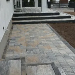Ułożona, szaro-beżowa kostka brukowa prowadząca do wejścia domu z trzema schodami, z widoczną instalacją klimatyzacji na ścianie i częściowo przygotowanym terenem pod trawnik.