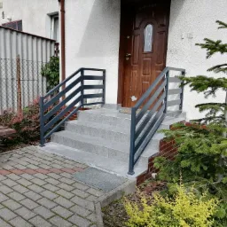 Szare, metalowe barierki schodowe prowadzące do brązowych drzwi wejściowych, z widocznym dywanikiem przed drzwiami i roślinnością po bokach.