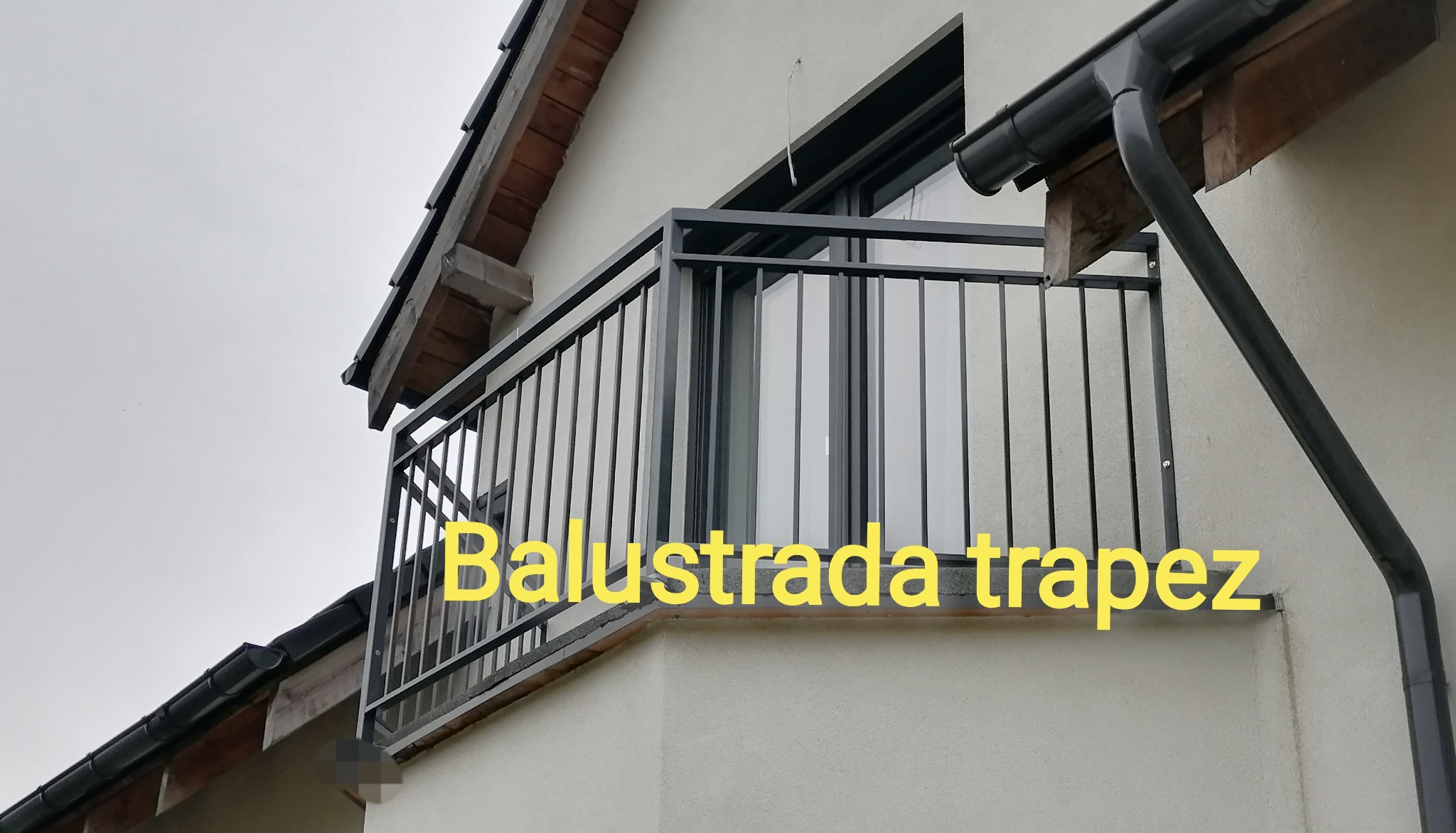 Balkon z metalową balustradą o prostym, nowoczesnym designie, widoczny z dołu pod kątem, z fragmentem rynny i dachu budynku w tle.