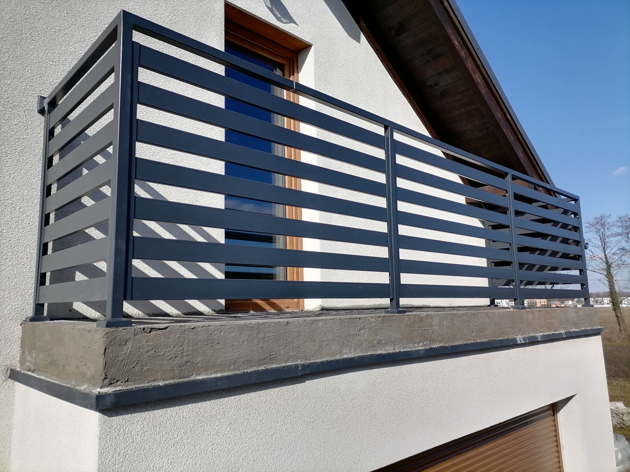 Balkon z nowoczesną, poziomą balustradą w kolorze grafitowym, zamontowaną na betonowej podstawie, z widokiem na okno i fragment dachu budynku.