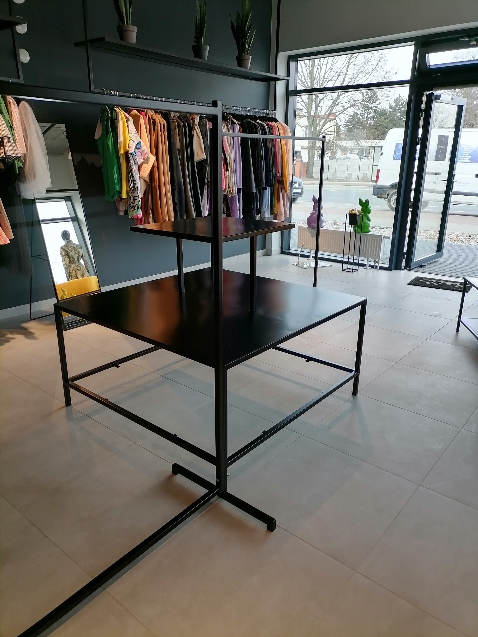 Czarny, metalowy regał sklepowy z trzema poziomami półek, widoczny na tle sklepu odzieżowego z wieszakami pełnymi ubrań i dużym lustrem