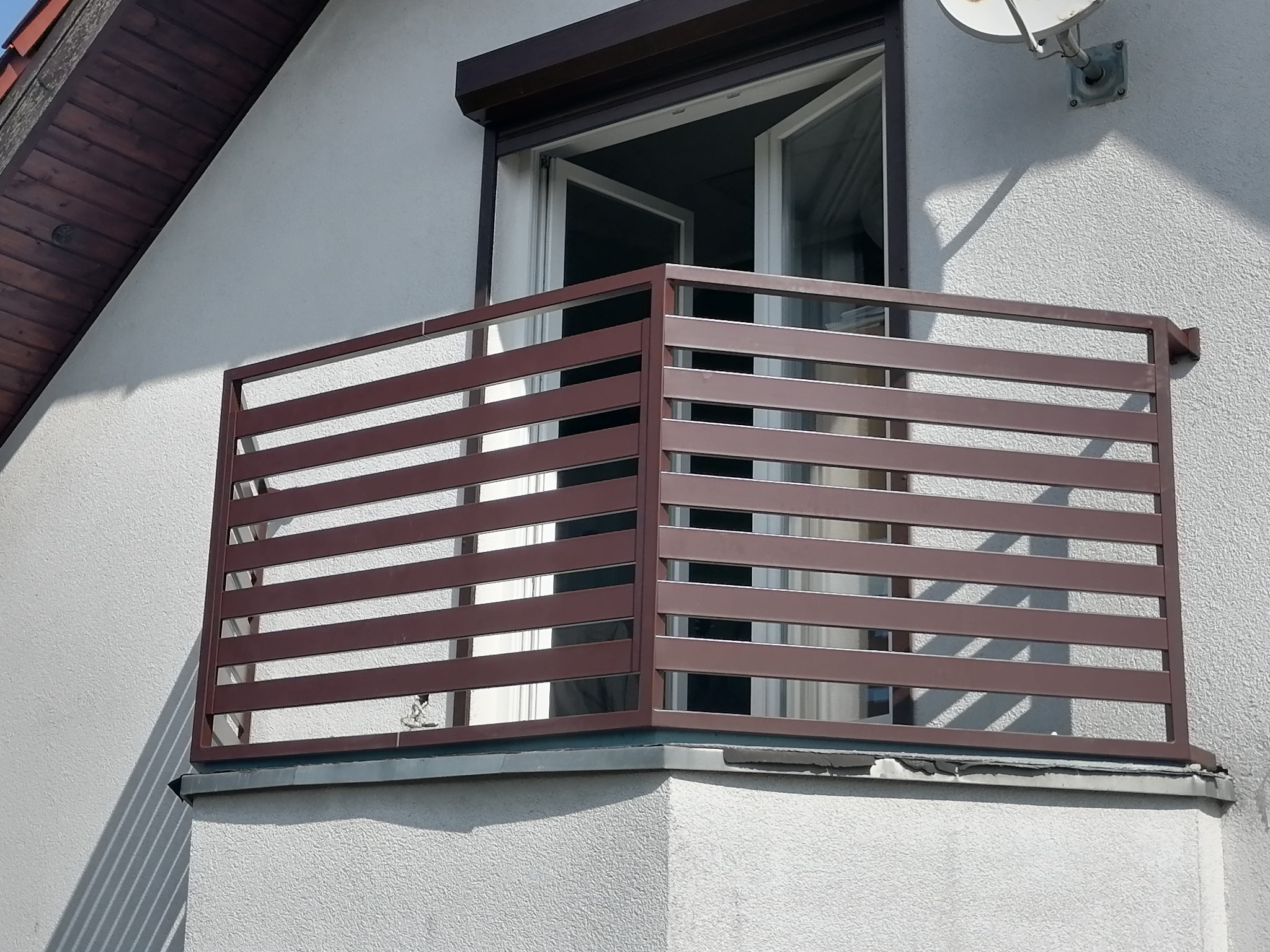 Brązowa, pozioma balustrada balkonowa z metalowych profili, zamontowana na balkonie z otwartym oknem i roletą zewnętrzną, widziana w słoneczny dzień.