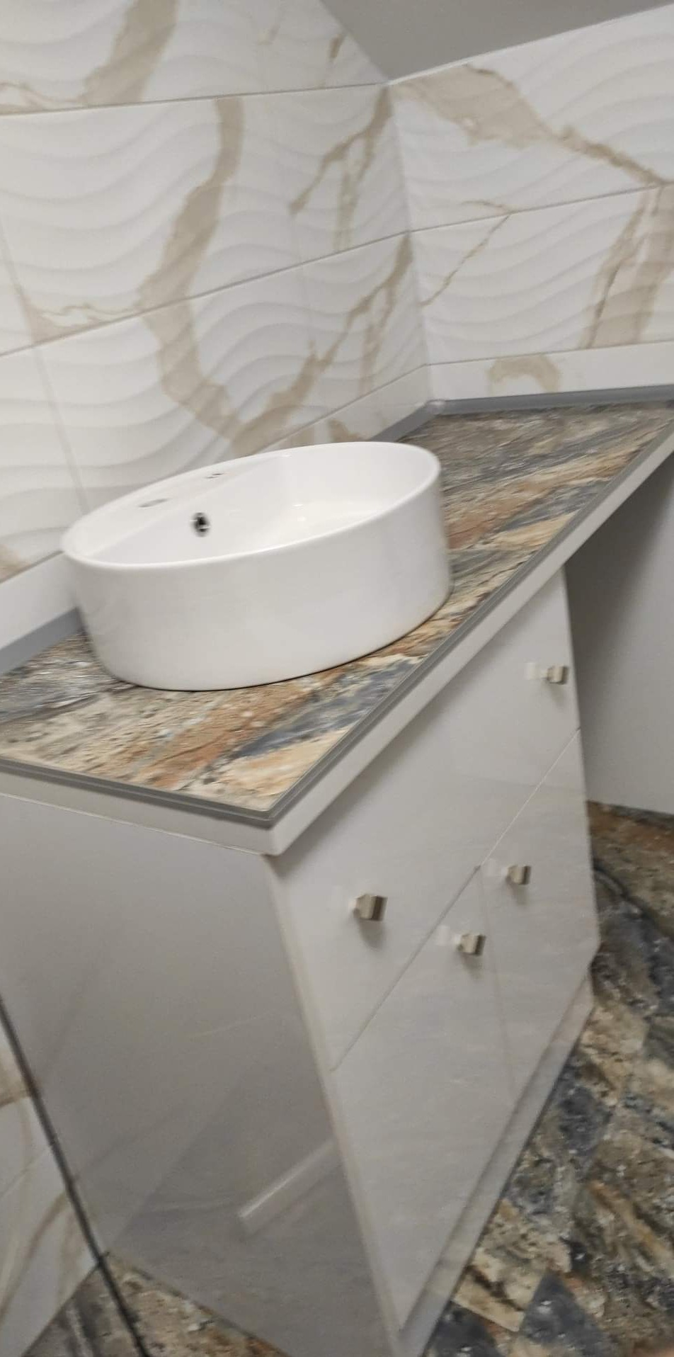 Biała, owalna umywalka nablatowa na szafce z czterema szufladami i blatem imitującym marmur, z płytkami ściennymi w tle o falistym wzorze.