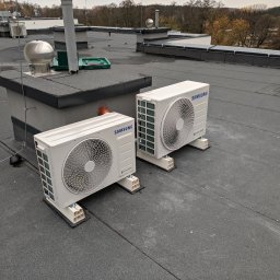 KANO TERM Karol Nowicki - Dwa białe agregaty klimatyzacyjne Samsung zainstalowane na płaskim, bitumicznym dachu budynku, widoczne inne elementy wentylacji dachowej i narzędzia w zielonej skrzynce.