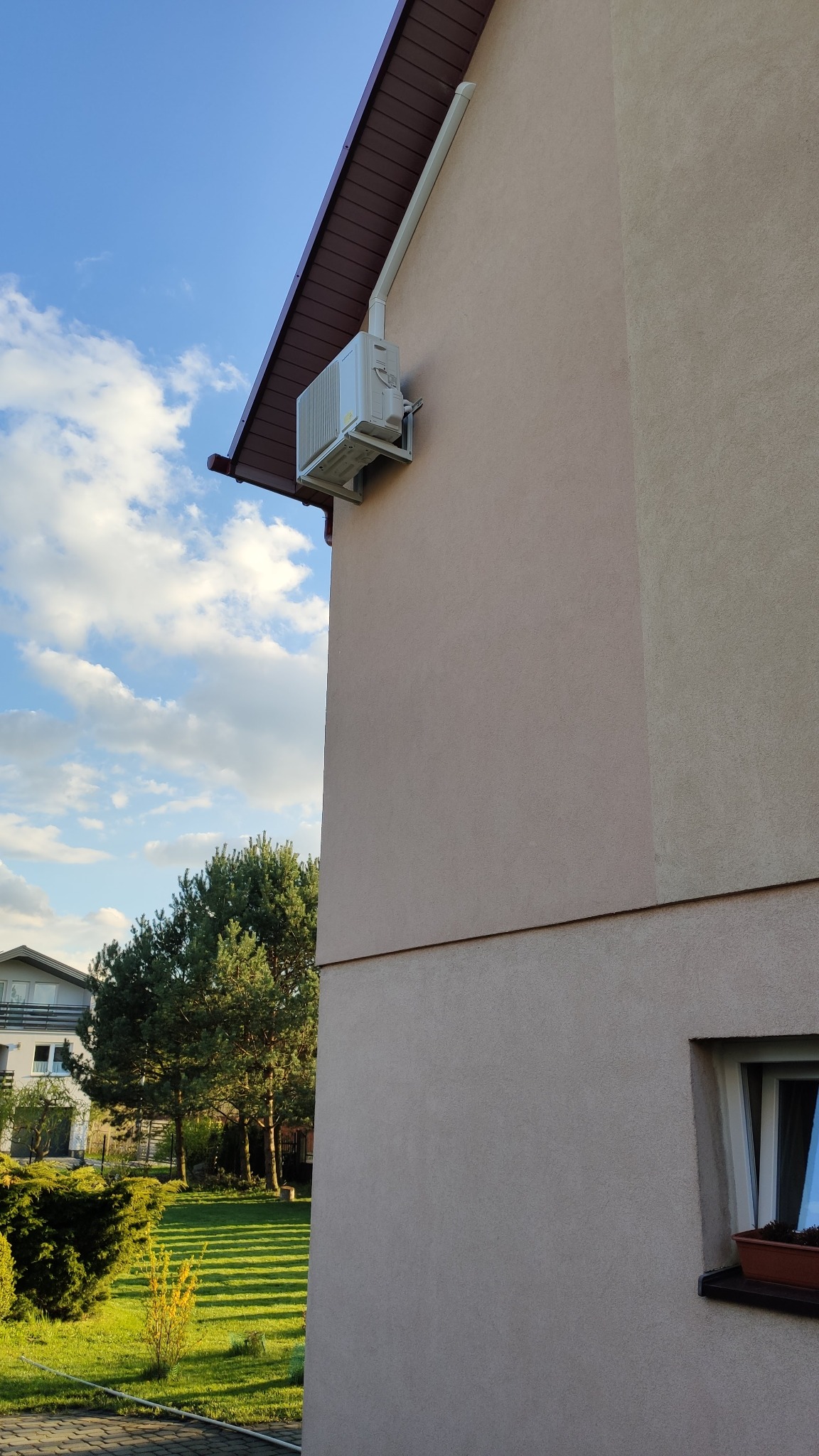 Zewnętrzny agregat klimatyzacji zamontowany na elewacji budynku z brązowym dachem, widoczny odpływ skroplin i fragment trawnika w tle.