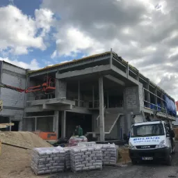 Budowa dwukondygnacyjnego budynku z betonu komórkowego, widoczny dźwig z pracownikiem na podnośniku, palety z materiałami budowlanymi oraz samochód dostawczy firmy Bolbud z numerem telefonu.