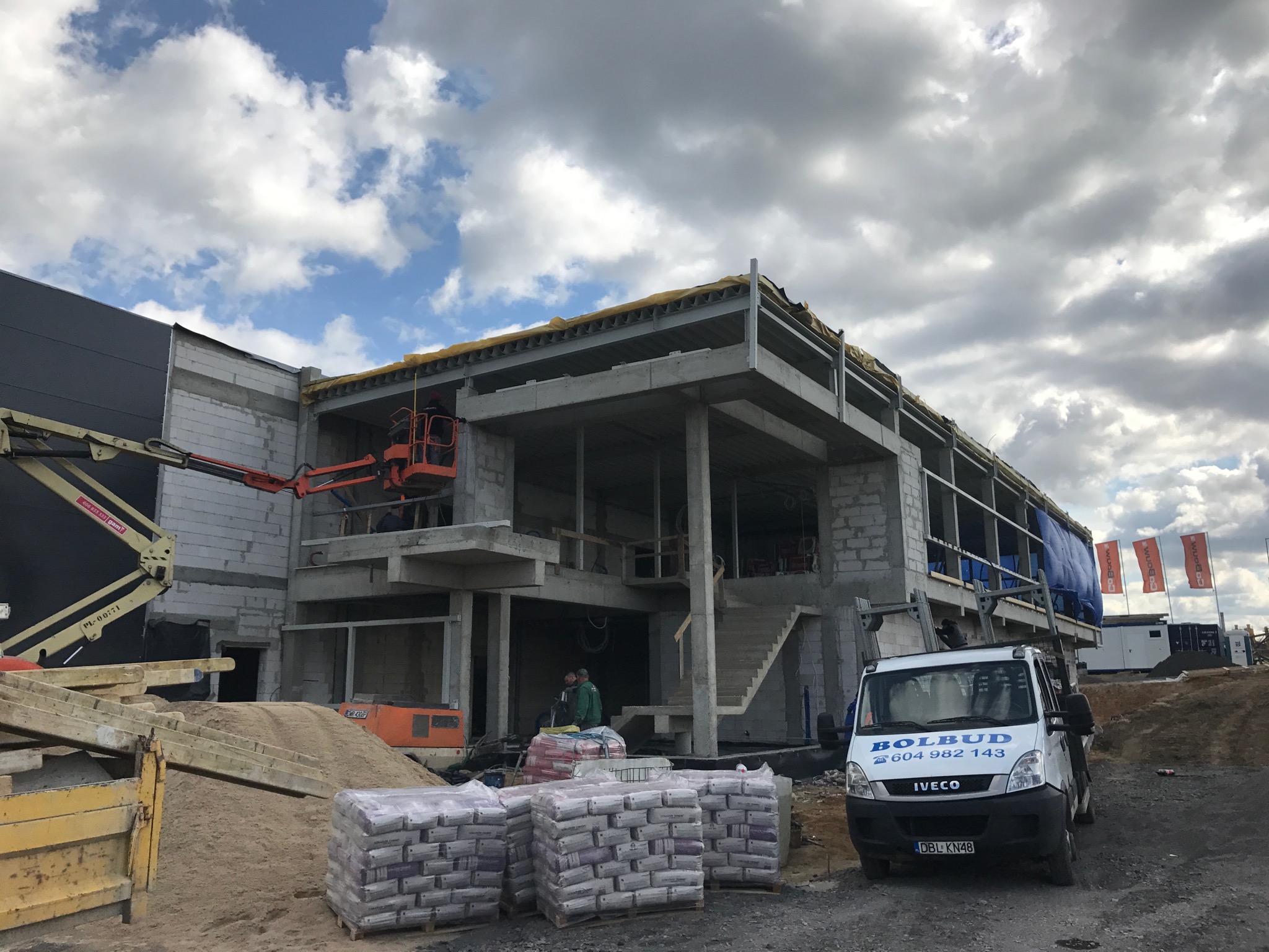 Budowa dwukondygnacyjnego budynku z betonu komórkowego, widoczny dźwig z pracownikiem na podnośniku, palety z materiałami budowlanymi oraz samochód dostawczy firmy Bolbud z numerem telefonu.