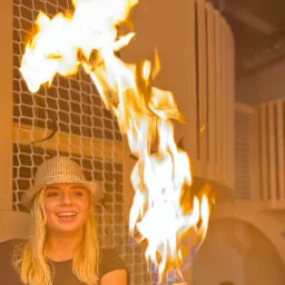 Uśmiechnięta blondynka w kapeluszu prezentuje płomienie na dłoni, w tle siatka i drewniana konstrukcja. Wyjątkowy pokaz iluzji ognia podczas eventu.
