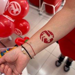 Wytwórnia Przyjęć - Brokatowy tatuaż z logo na ramieniu kobiety, w tle czerwone balony z tym samym logo. Dekoracja imprezowa.