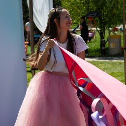 Wytwórnia Przyjęć - Uśmiechnięta animatorka w różowej tiulowej spódnicy trzyma kolorową girlandę na tle zieleni i namiotu. Słoneczny dzień, impreza plenerowa.