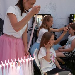Wytwórnia Przyjęć - Animatorzy malują twarze dzieciom i zaplatają kolorowe warkoczyki podczas imprezy plenerowej pod namiotem z motywem jednorożca. Widoczne akcesoria do stylizacji.