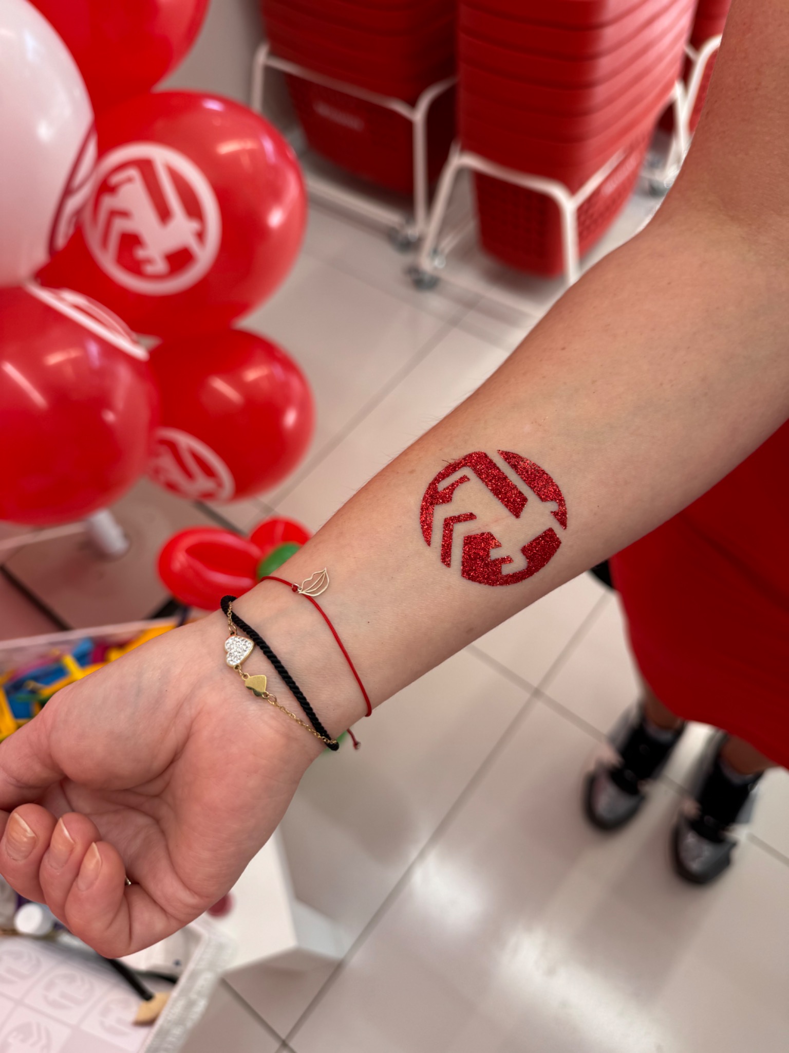 Brokatowy tatuaż z logo na ramieniu kobiety, w tle czerwone balony z tym samym logo. Dekoracja imprezowa.