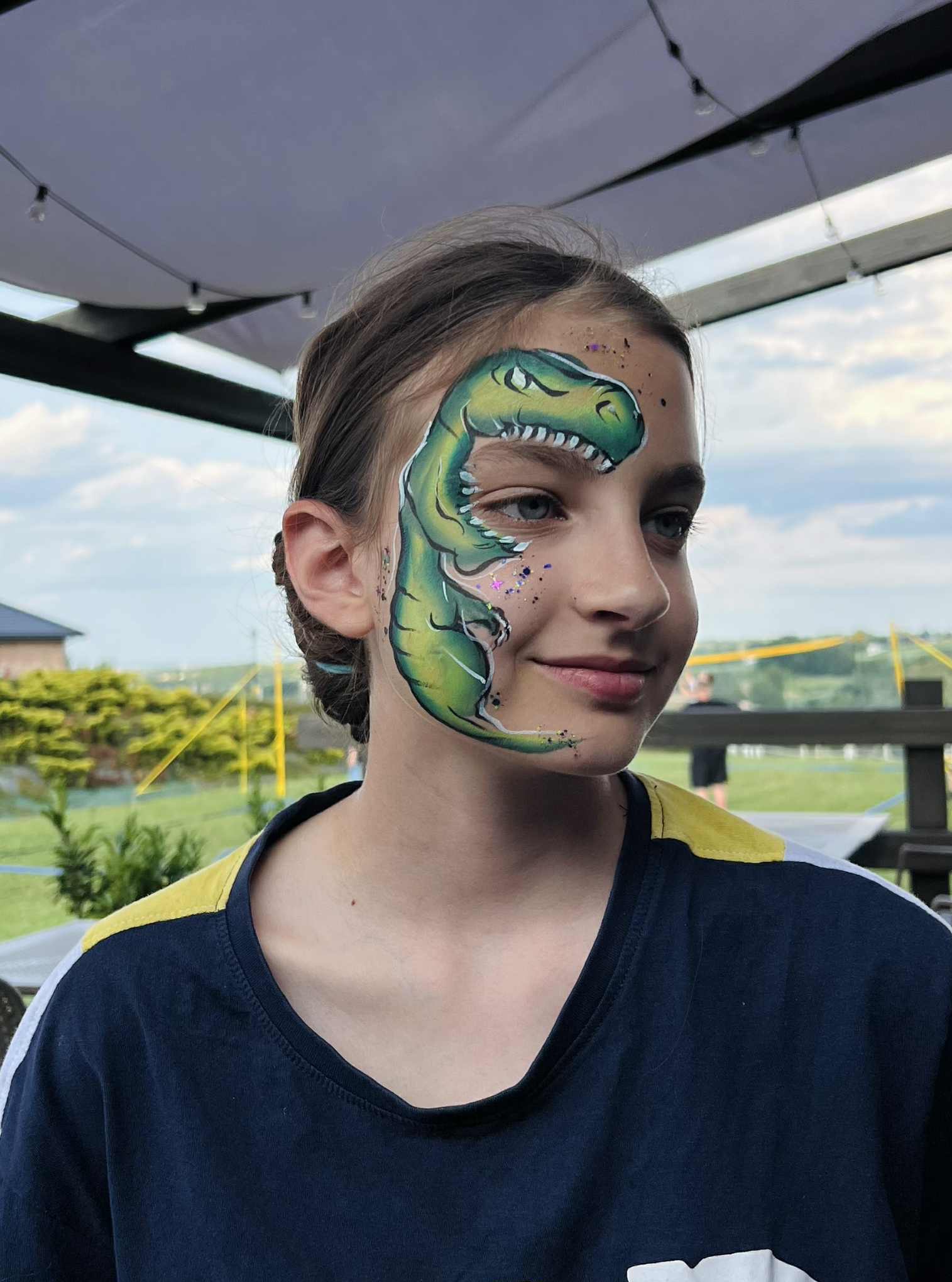 Dziewczynka z pomalowaną twarzą w dinozaura, z brokatem, na tle ogrodu i białego zadaszenia. Uśmiechnięta, z profilu.