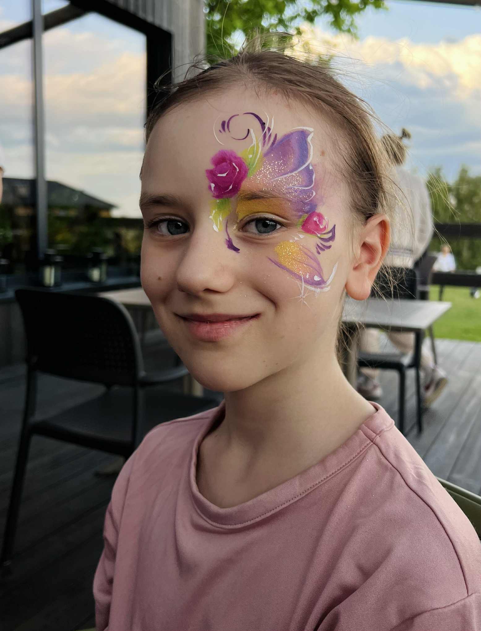 Uśmiechnięta dziewczynka z profesjonalnym, artystycznym makijażem na twarzy w motywy kwiatowe i motyla, wykonanym farbami do ciała, na tle plenerowym.