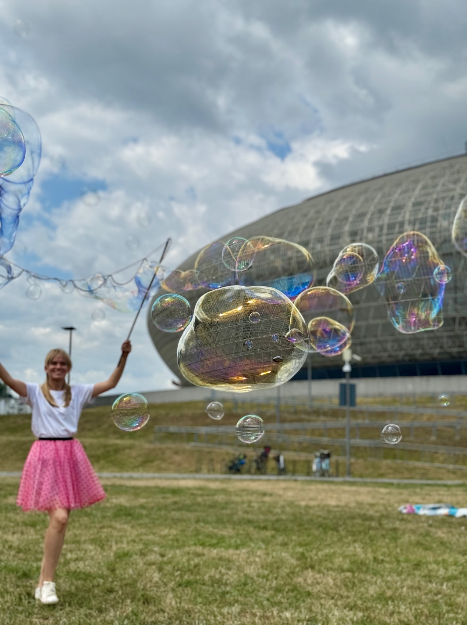 Uśmiechnięta kobieta w różowej spódnicy puszcza duże bańki mydlane na trawniku. W tle widoczny budynek o siatkowej konstrukcji i błękitne niebo z chmurami.