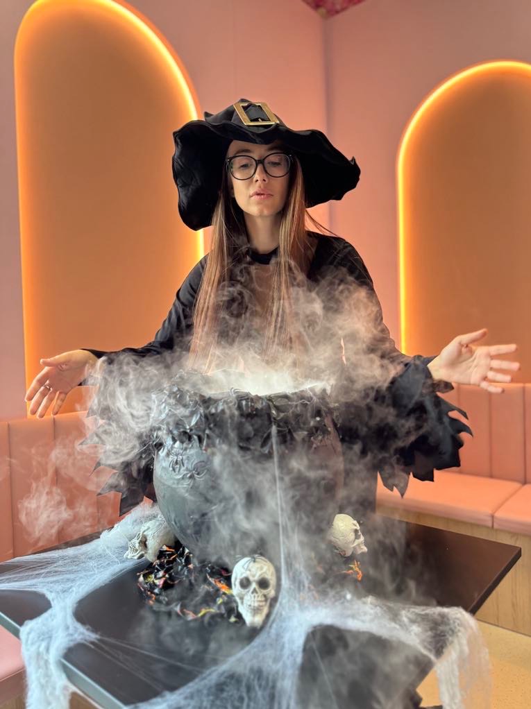 Kobieta w kostiumie wiedźmy, z okularami, pochyla się nad dymiącym kotłem, dekoracje halloweenowe, wnętrze z pomarańczowym oświetleniem.