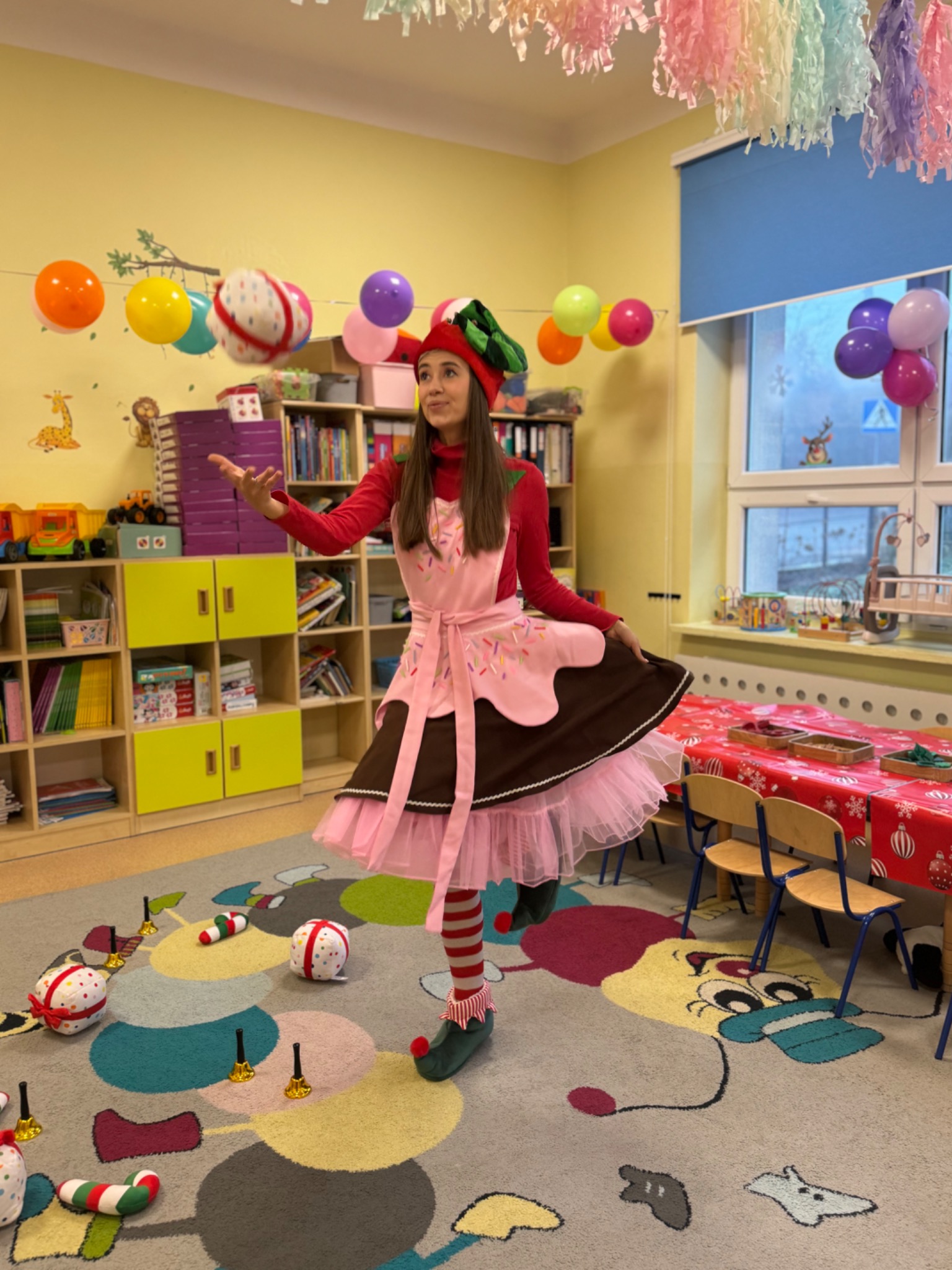 Animator w kostiumie cukiernika żongluje piłkami w udekorowanym pokoju dziecięcym. Kolorowe balony i dekoracje tworzą radosną atmosferę imprezy.