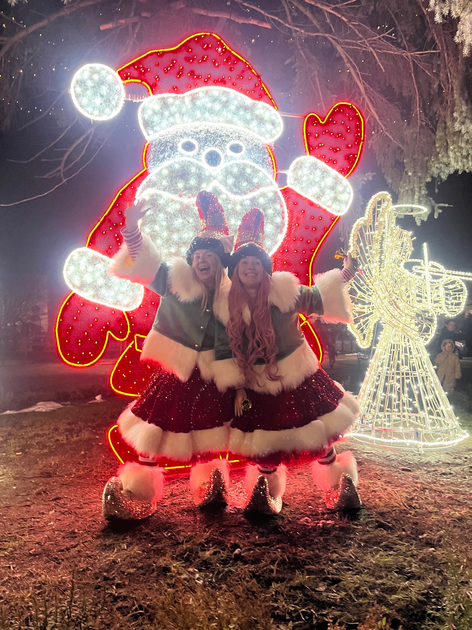 Dwie uśmiechnięte kobiety w kostiumach elfów na tle świetlnej dekoracji świątecznej z motywem Mikołaja i anioła, w scenerii zimowej nocy.