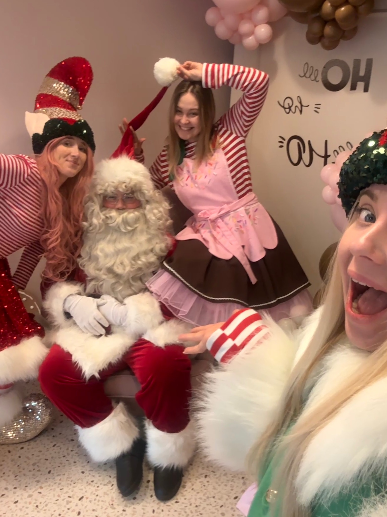 Wesołe selfie z Mikołajem i elfami w paski, dekoracje świąteczne w tle. Uśmiechnięte twarze i radosna atmosfera imprezy bożonarodzeniowej.
